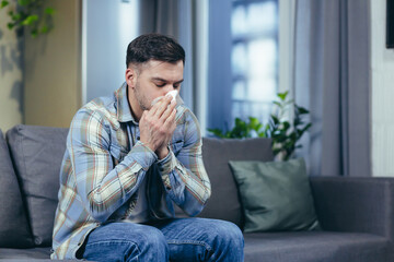 Fototapeta premium Feeling unwell. A young handsome man is sitting on the couch at home, is sick, feels cold, wipes his nose with a napkin, has a runny nose.