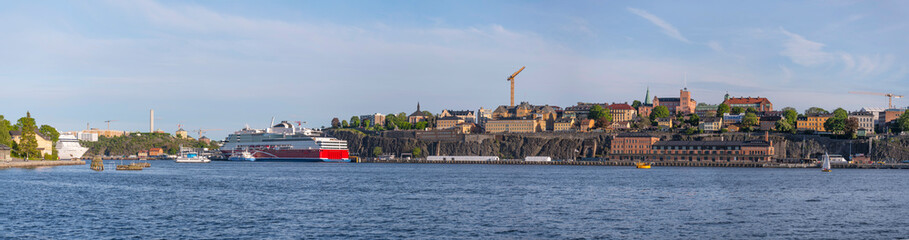 Obraz premium Panorama view over the bay Stockholms Ström in evening light with the district Södermalm and cruise ships and island with a castle and brick building a sunny evening in Stockholm