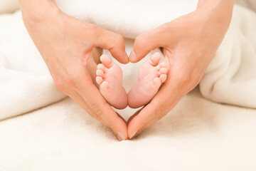 Newborn cute barefoot in heart shape created from young adult mother hands on white soft blanket in bed. Lovely emotional, sentimental moment. Closeup. Parent love. Front view.