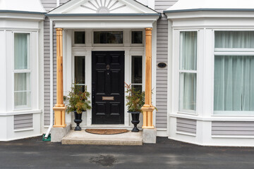 A black metal formal looking door with two flower pots on both sides. The entrance to the grey house has glass windows and wood columns. The siding is a wood clapboard with white trim. 