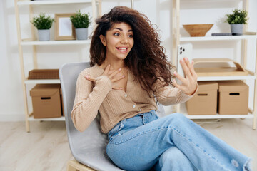 Excited happy cute tanned curly Latin lady doing selfie video call with trendy smartphone sit on chair in home modern interior look at screen. Copy space Mockup Banner. Concept social network