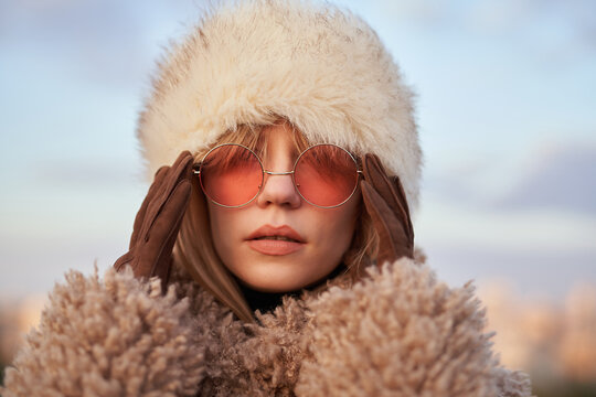Attractive glamour young woman in stylish clothes - artificial fur coat and hat correcting pink sunglasses with hands in brown gloves on background of blue cloudy sky. High quality image