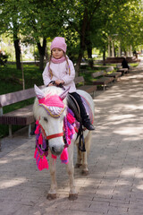 Obraz premium Little girl riding a pony horse in park. Child dressed in pink jacket and hat on horse.