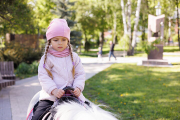 Little girl riding a pony horse in park. Child dressed in pink jacket and hat on horse.