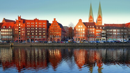 Obraz premium historische Häuserfassaden und Kirchtürme in Lübeck spiegeln sich entlang der Trave im Abendlicht
