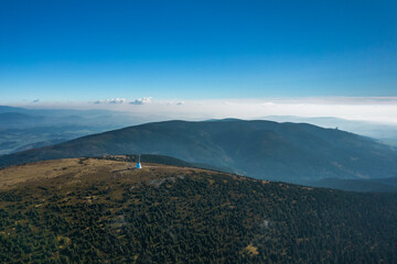 Aerial landscape of the green mountains and coniferous forest in a sunny day. Blue sky and white clouds over the peaks. View of the tower observation in the middle.