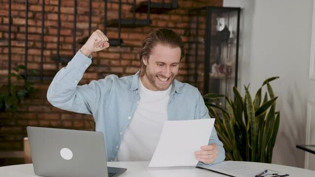 Overjoyed employee shows happines ove clenching fists looking in some documents or reports. Successful young entepreneur.
