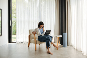 Relaxed young caucasian girl sits on armchair drinks morning coffee and uses digital tablet. Brunette wears casual clothes relaxing at home. Social media addiction concept