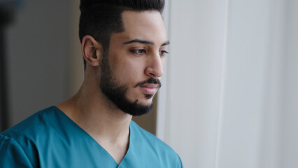 Young pensive hispanic doctor male thoughtful arabian nurse practitioner therapist surgeon stand near window in hospital workplace wait for patient visit sad medical man think about ending quarantine