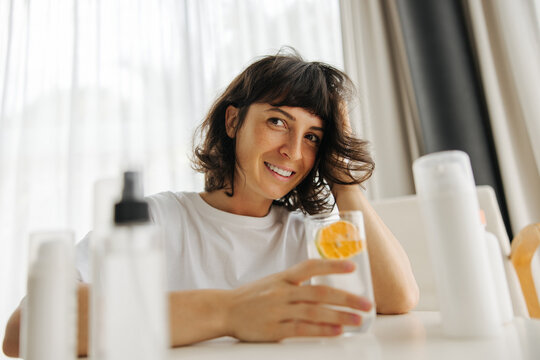 Attractive young caucasian woman with snow-white smile drinks water sitting at table. Brunette with short haircut wears white t-shirt. Lifestyle concept