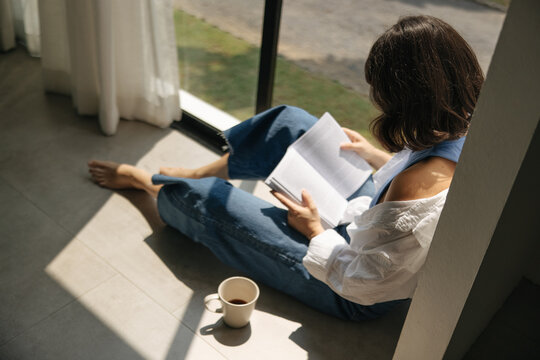 Top view of young caucasian woman reading book sitting by window in sunny weather. Brunette in casual clothes starts morning with cup of coffee. Lifestyle concept