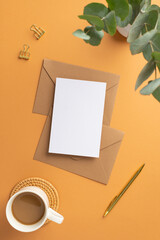 Top view vertical photo of workspace paper sheet over craft paper envelopes gold pen clips cup of coffee on rattan serving mat and vase with eucalyptus on isolated orange background with blank space
