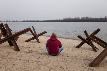 Ukrainian civilian man sitting on river bank among anti tank czech hedgehogs. Russian war in Ukraine 2022. Protecting freedom from terrorism © elenaseiryk