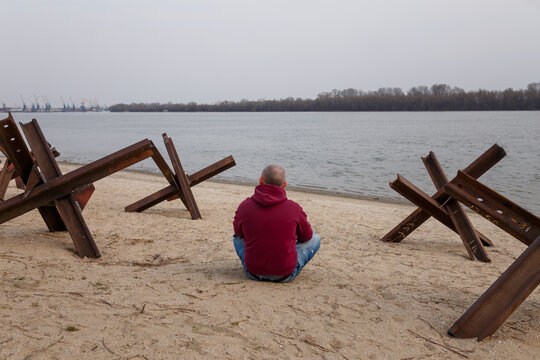 Ukrainian civilian man sitting on river bank among anti tank czech hedgehogs. Russian war in Ukraine 2022. Protecting freedom from terrorism