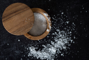 Top view of sea salt on dark moody background inside round wooden salt container.