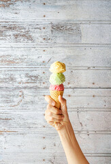 Woman's hand holding wafer cone with strawberry, pistachio and vanilla ice cream. Close-up, wooden background.