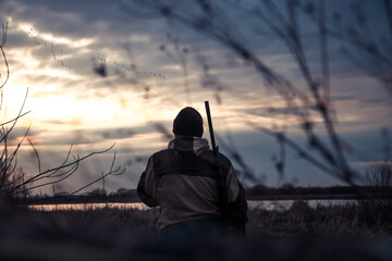 Hunter man in camouflage with shotgun looking into the distance with flying gooses on horizon during dramatic sunset during hunting season  - obrazy, fototapety, plakaty