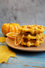 Belgian gluten- and lactose-free waffles with pumpkin stand on a gray plate with a small pumpkin and dry lemon chips on a gray table with yellow maple leaves. side view