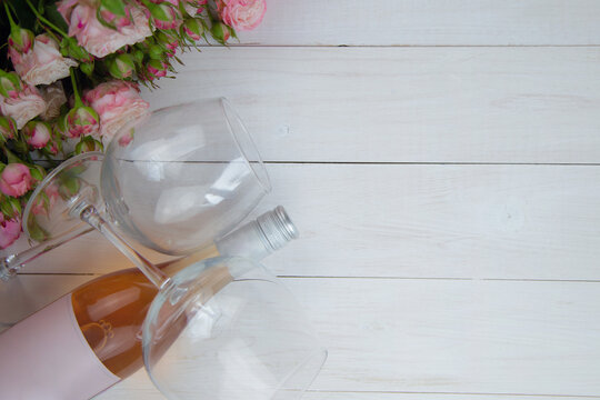 A large bouquet of pink roses and a bottle of rose wine with glasses lie on the side on a wooden white background with a place for text. High quality photo