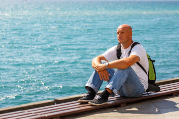 Pensive handsome middle-aged man traveler with backpack in white t-shirt and blue jeans, sits and...