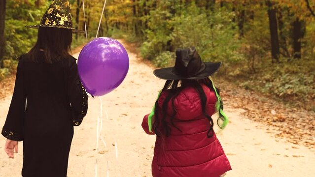 Back view video of mom holding the violet balloon and her daughter in suits for Halloween walking on the path in the forest.