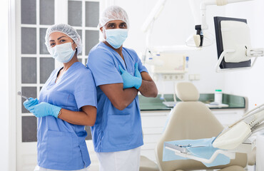 Obraz premium Two professional hispanic dentists in blue uniforms standing back to back in modern office of dental clinic