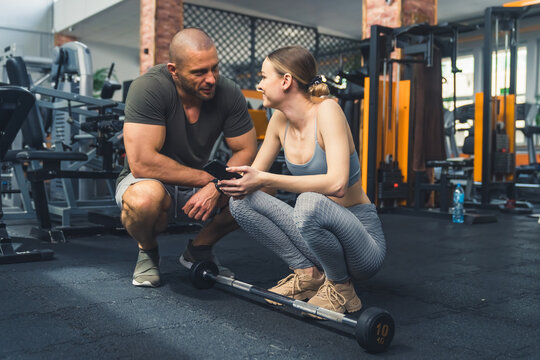 Indoor shot at a gym of two caucasian people. Woman in a sports outfit crunching on the floor with a barbell in front of her, man crunching near her talking to her. High quality photo