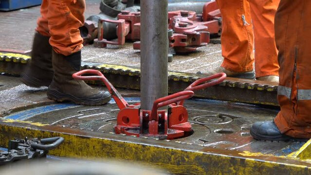 Drilling platform. Rotor table. Manual slips installed. Legs in orange coveralls. Drill floor. RIG.