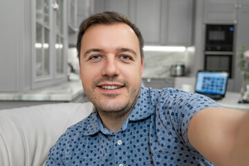 Image of millennial man smiling and taking selfie photo while sitting on sofa at home. Portrait of happy man in blue shirt taking selfie or having video call with friends showing new flat