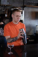 stylish young man hipster in orange t-shirt making mixing a cocktail in a dark loft cafe. alcohol drink in modern bar