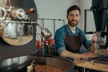 Smiling barista drinking coffee sitting near coffee roasting machine