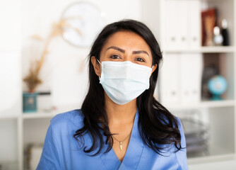 Latin American female nurse wearing face mask while writing notes and reading something on clipboard at her office during coronavirus quarantine