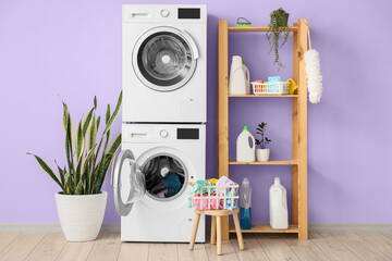 Modern drying, washing machines, basket with laundry and shelf unit near violet wall