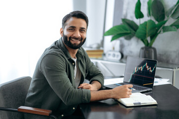 Portrait of a handsome attractive positive successful Indian or Arabic crypto investor, stock exchange broker, cryptocurrency expert, sit at a desk in modern office, looks at camera, smiling friendly