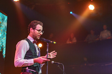 Attractive and confident Public speaker with glasses bow tiea a mobile phone in hand in the spotlight talking in conference or concert hall. People blurred in background