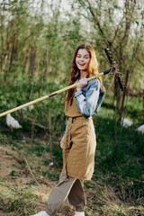 A woman farmer in work clothes and an apron works outdoors in nature and holds a rake to gather grass