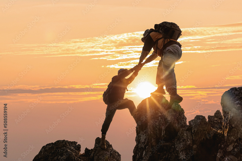 Wall mural silhouette two male hikers climbing up mountain cliff and one of them giving helping hand. people he