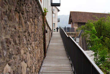 Fototapeta na wymiar Pathway along a stone wall to wooden covered bridge at Village of Stein, Canton Aargau, on a cloudy spring day. Photo taken May 6th, 2022, Stein, Switzerland.