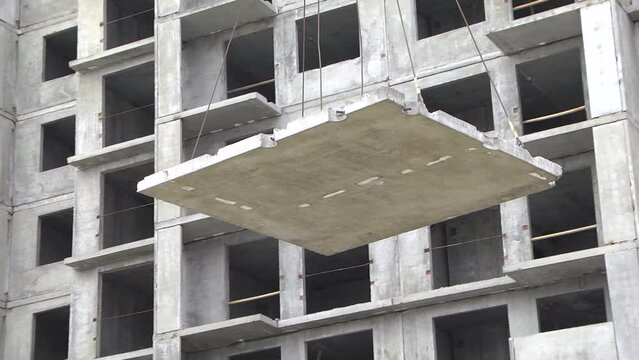 Concrete building and tower crane's hook over it in the construction site. Concrete slab hanging from crane hook above building skeleton at construction site
