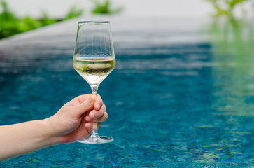 Hand holding a glass of white wine with swimming pool background. Holiday and summer drink concept.