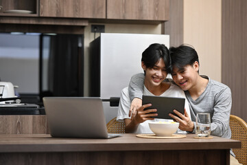Happy gay couple embracing and surfing internet with digital tablet together at kitchen table. LGBT, pride, relationships and equality concept