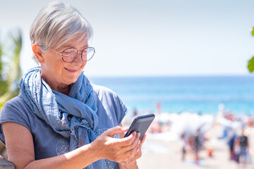 Smiling white haired senior woman reading messages on smart phone outdoor on a sunny day near the...