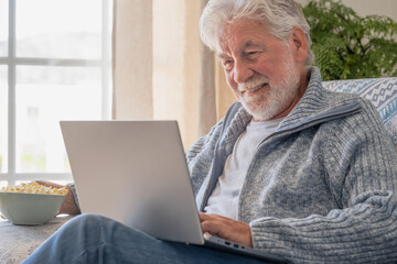 Elderly 70s man seated on sofa browsing on laptop, senior looking at laptop screen surfing the net, older generation and modern tech application easy usage concept