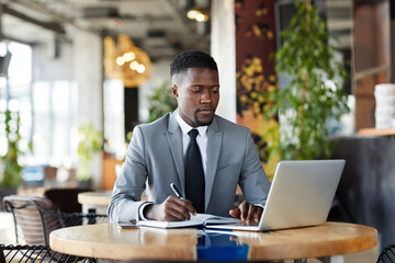 Serious busy young African-American entrepreneur in gray suit sitting at table and planning tasks, he making notes in diary while using laptop in cafe