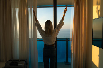 Young woman in a white shirt and blue jeans looks at the sea standing with her hands up at the window in a hotel room. Concept of travel and summer holidays