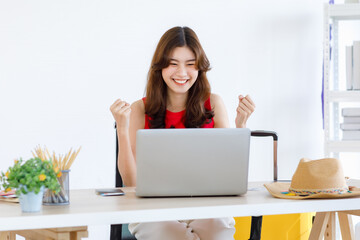 Asian excited happy female traveler in casual outfit sitting looking at laptop computer holding...