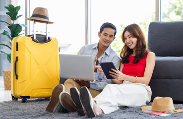 Asian young lovely boyfriend and girlfriend traveler couple sitting smiling on carpet in living room with trolley luggage while using laptop computer and tablet booking vacation summer trip online