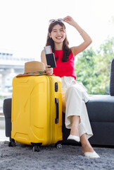 Asian young happy cheerful female traveler in casual summer outfit with sunglasses sitting smiling on cozy sofa in living room holding passport and boarding pass ticket with yellow trolley luggage