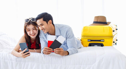 Asian young traveler lover couple in casual outfit with sunglasses and hat laying down smiling together on bed holding passport boarding pass ticket using smartphone booking hotel reservation online