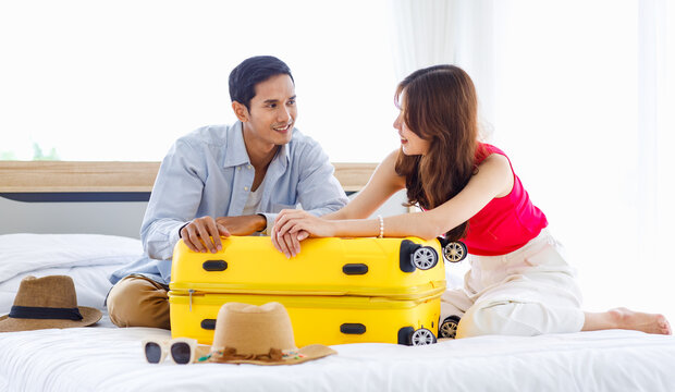 Asian young romantic lover couple boyfriend and girlfriend in casual outfit sitting smiling helping packing luggage together on bed while male using tablet computer booking vacation trip hotel online